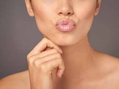 A person with a neutral skin tone puckers their lips, enhanced with lip filler, wearing light pink lipstick, and gently places fingers on their chin. The background is a plain, neutral color.